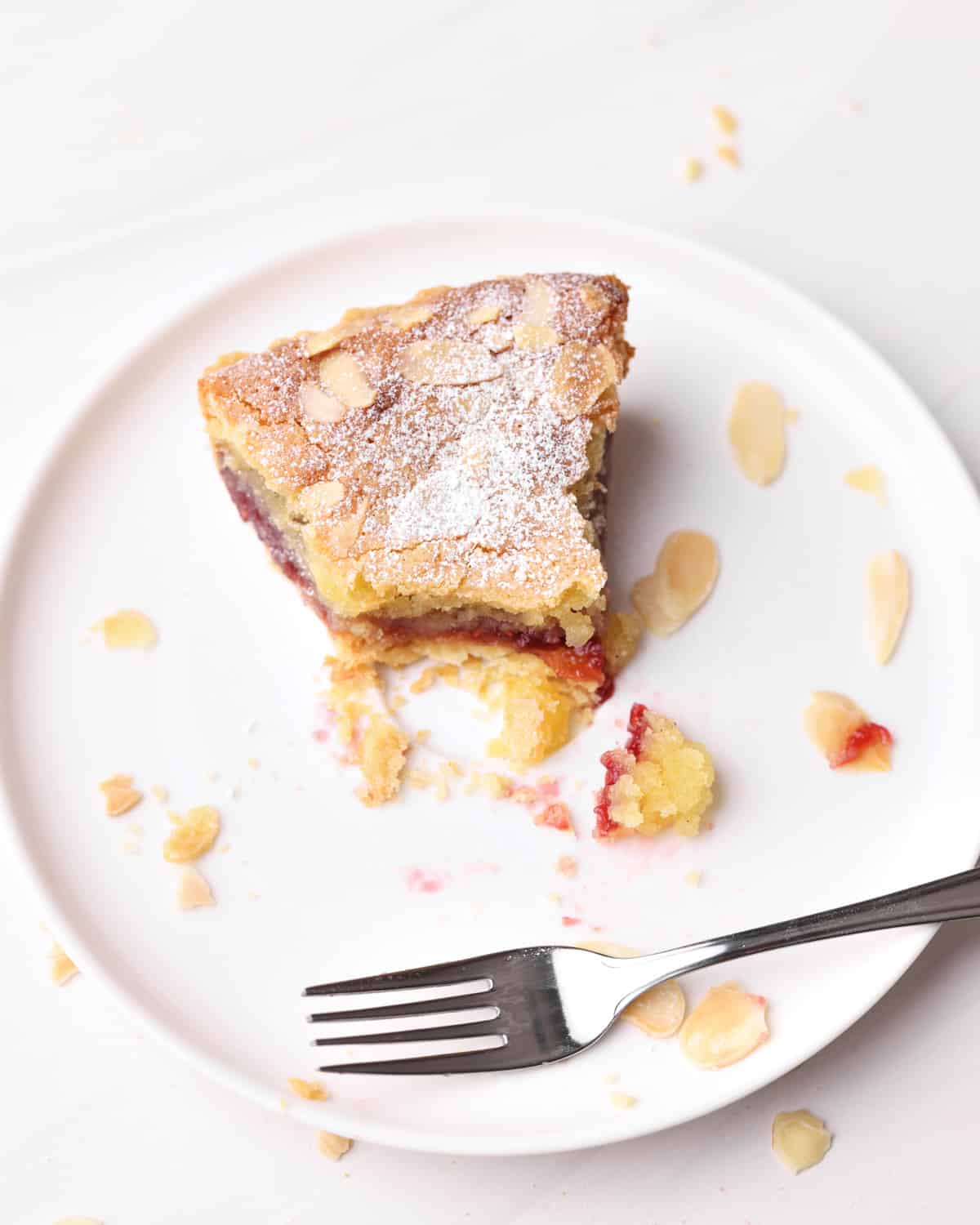 Slice of bakewell tart with a bite taken out of it, on a plate with a fork. 