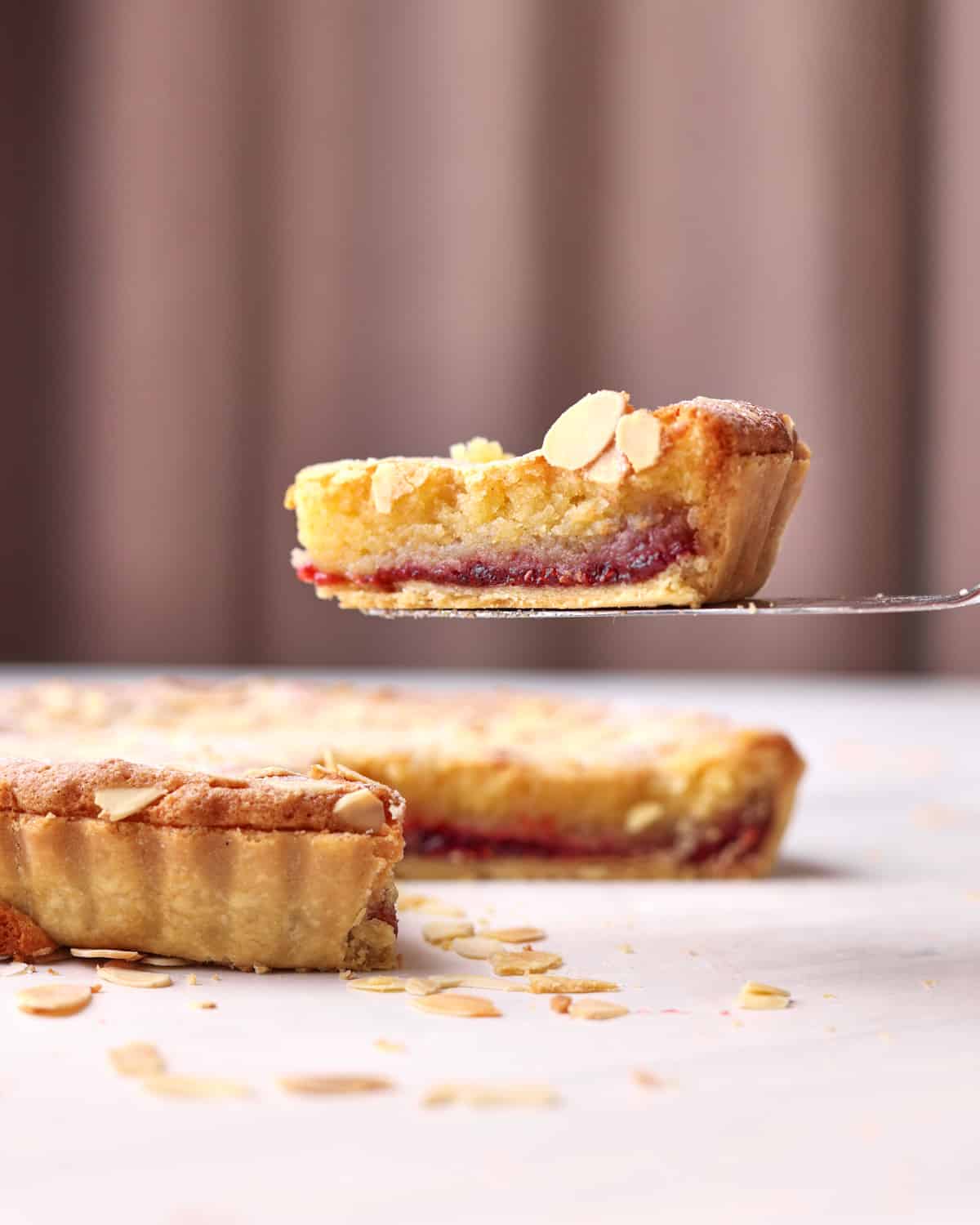 Side view of a bakewell tart with a slice being lifted out of it. 