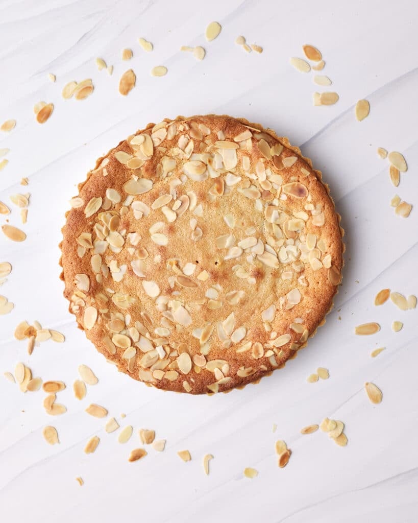 A baked bakewell tart, fresh out of the oven, on a white background surrounded by toasted flaked almonds. 