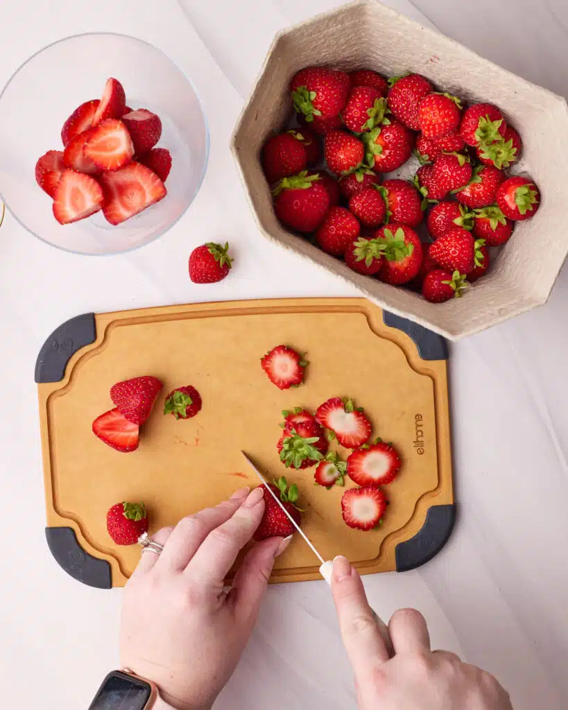 Cutting the tops off of and slicing strawberries on a small chopping board. 