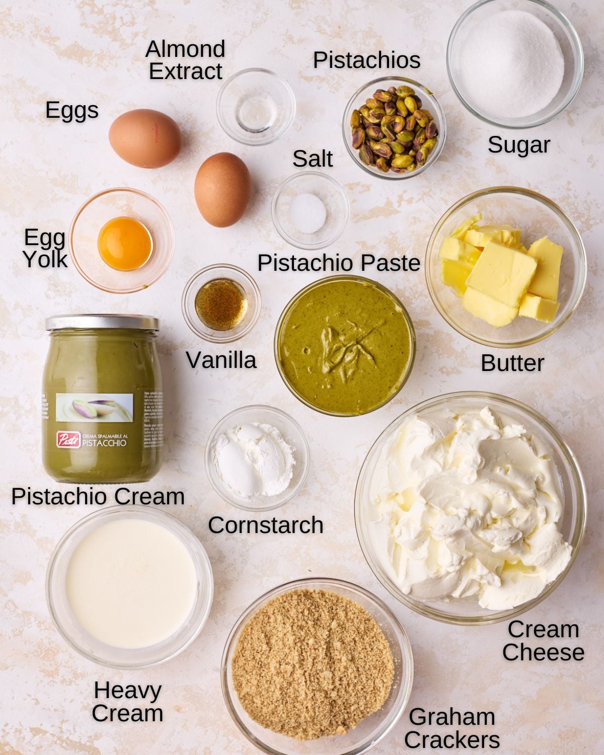 Ingredients to make pistachio cheesecake - Cream cheese, sugar, eggs, cornstarch, egg yolk, graham cracker crumbs, heavy cream, butter, vanilla, almond extract, salt, pistachios, pistachio paste and pistachio cream.