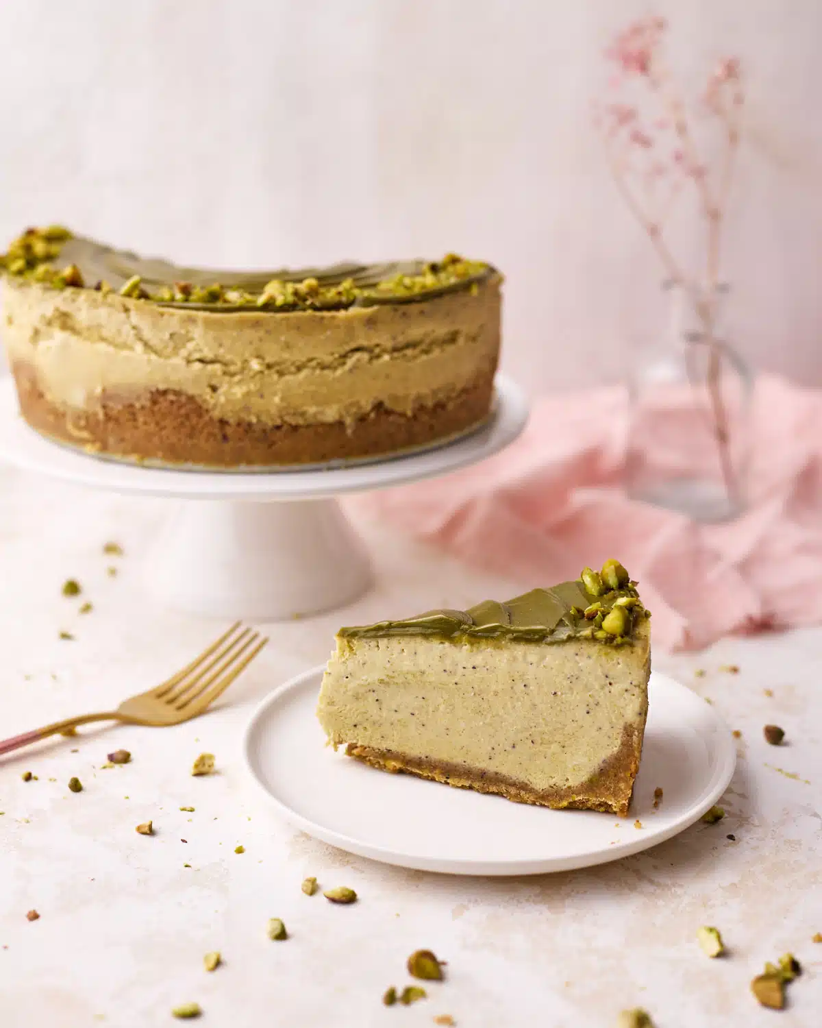 Pistachio cheesecake on a cake stand with one slice on a plate in the foreground - pink napkin in the background. 