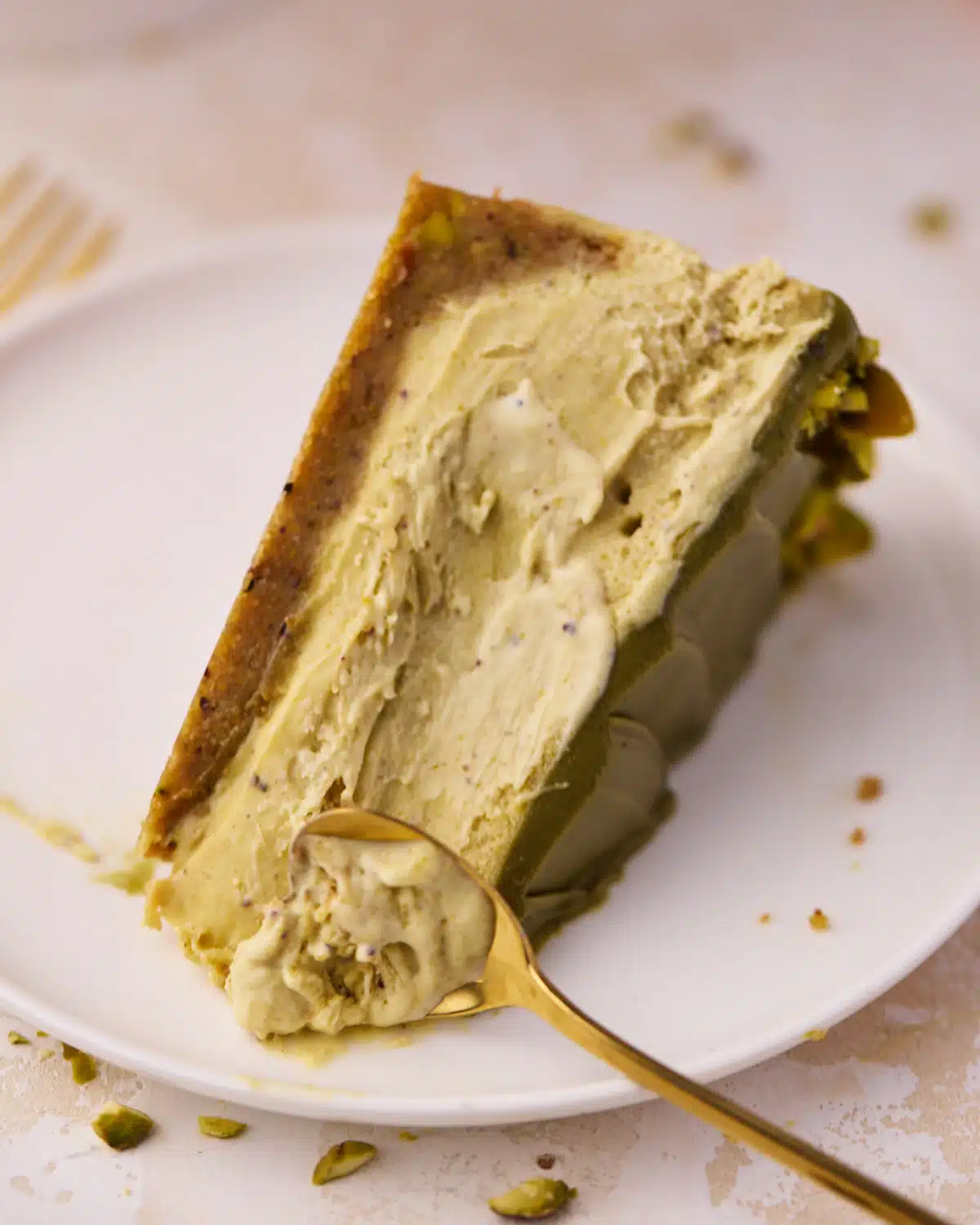 Slice of pistachio cheesecake on its side, a small spoon is being run down the centre to scoop it out and show how soft and creamy it is. 