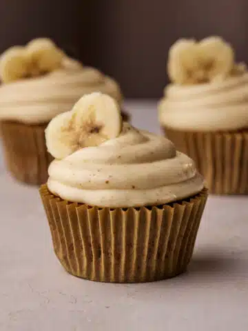 Three banana cupcakes with brown butter cream cheese frosting and a heart shaped banana slice on top.