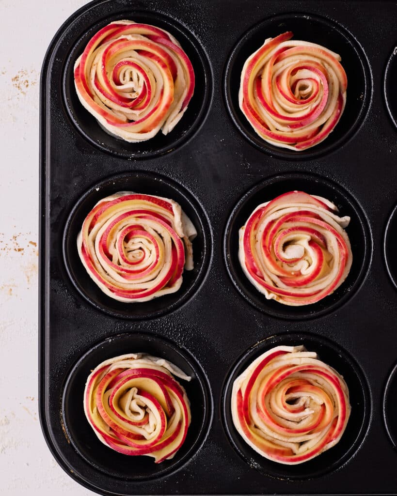 Six apple roses sitting inside the cavities of a muffin pan, ready to be baked.