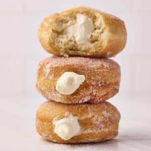 Three Bavarian cream donuts sitting in a stack, the top one has a bite taken out of it to reveal the creamy filling.