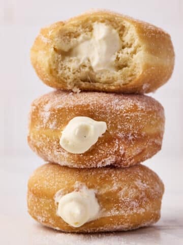 Three Bavarian cream donuts sitting in a stack, the top one has a bite taken out of it to reveal the creamy filling.