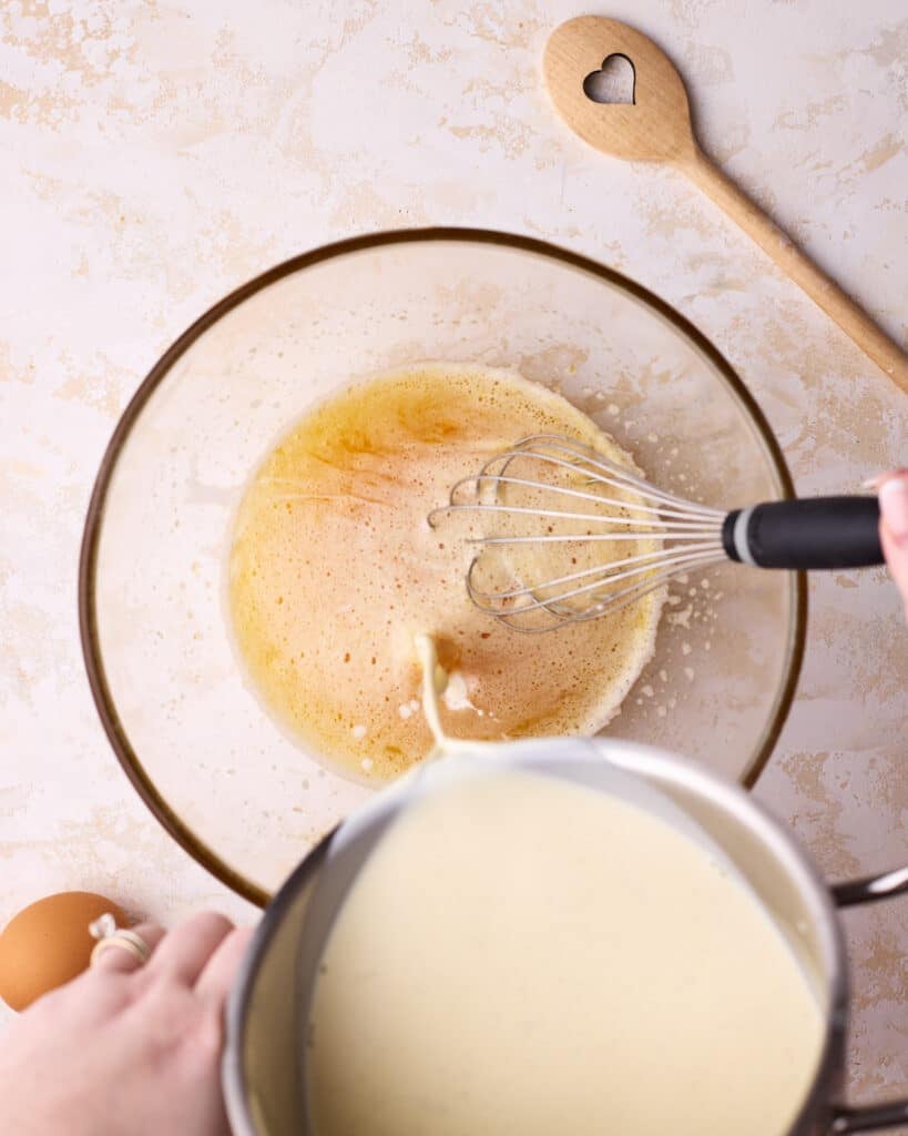 Drizzling hot milk mixture from a saucepan into a bowl of whisked eggs and sugar to gradually temper the eggs for custard pie filling. 