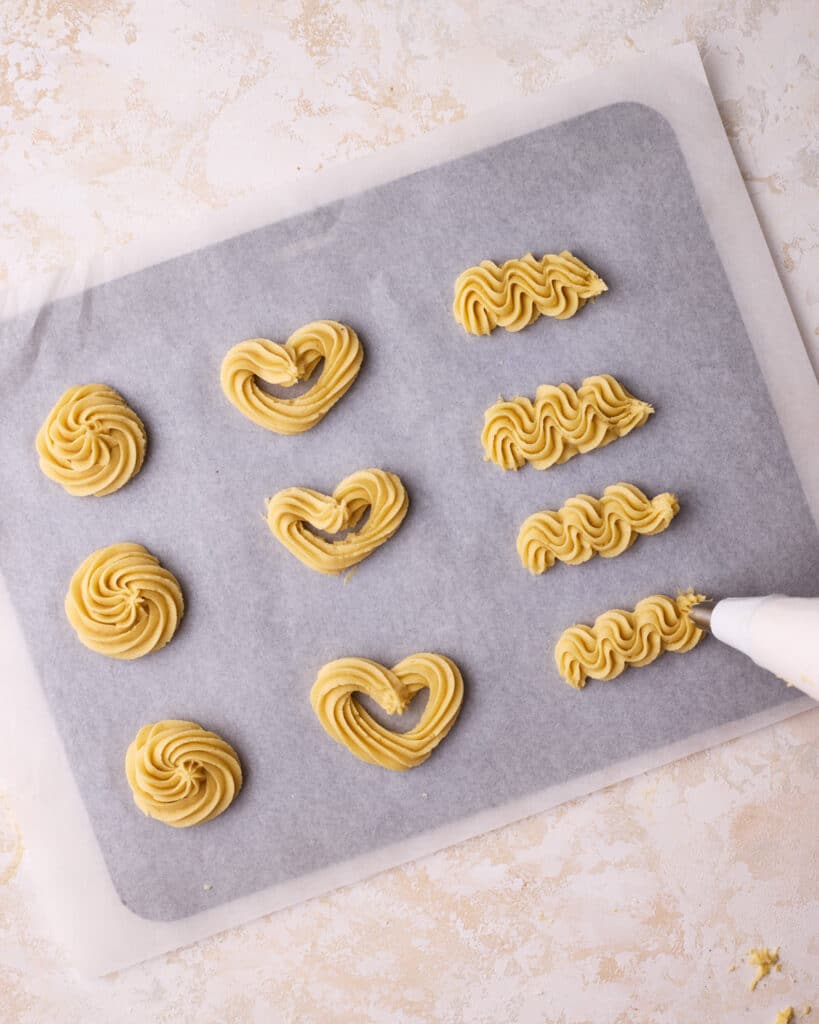 Butter cookie dough being piped into various shapes on a baking sheet to make Danish butter cookies. 