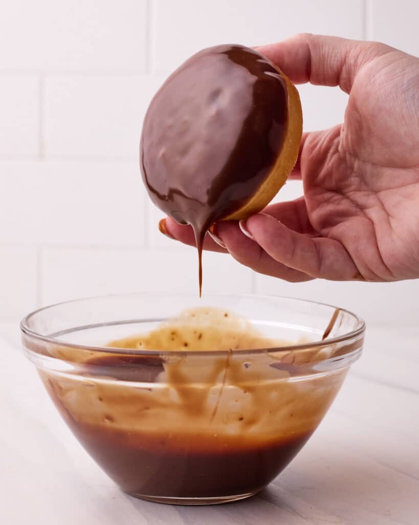 Dipping a boston cream donut into a small glass bowl filled with chocolate ganache glaze. 