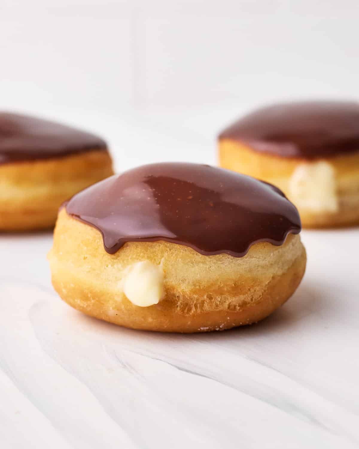 Three boston cream donuts on a table with a little pastry cream spilling out the front and a shiny chocolate glaze on top. 