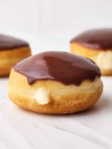 Three boston cream donuts on a table with a little pastry cream spilling out the front and a shiny chocolate glaze on top.