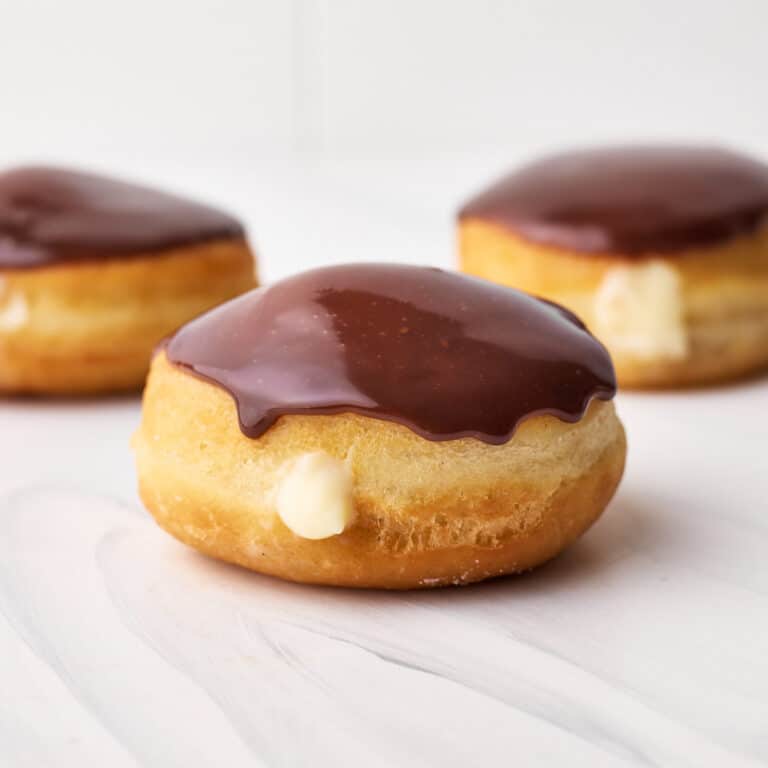 Three boston cream donuts on a table with a little pastry cream spilling out the front and a shiny chocolate glaze on top.