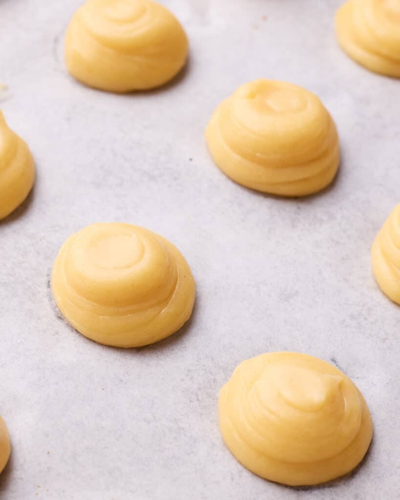 Little domes of choux pastry on a baking sheet with the peaks flattened to make profiteroles. 