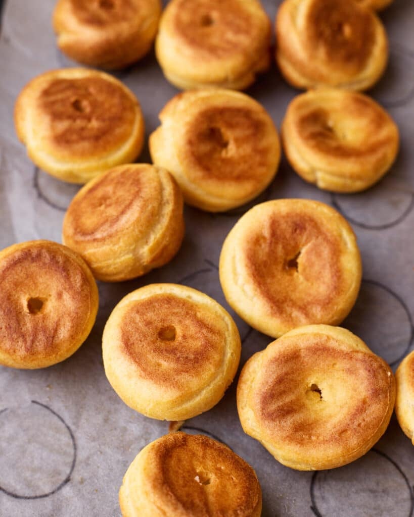 Upside down profiteroles on a baking sheet, showing little holes pierced in the bottom to let steam release. 