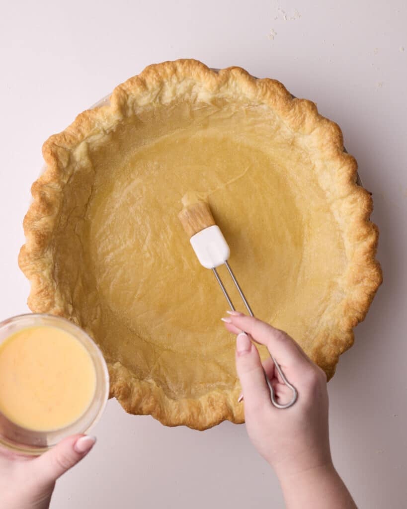 Brushing egg wash onto a partially baked pie crust with a pastry brush.