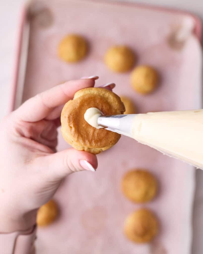 One profiterole being held while a pastry bag of creamy filling is being squeezed into it through the hole in the bottom. 