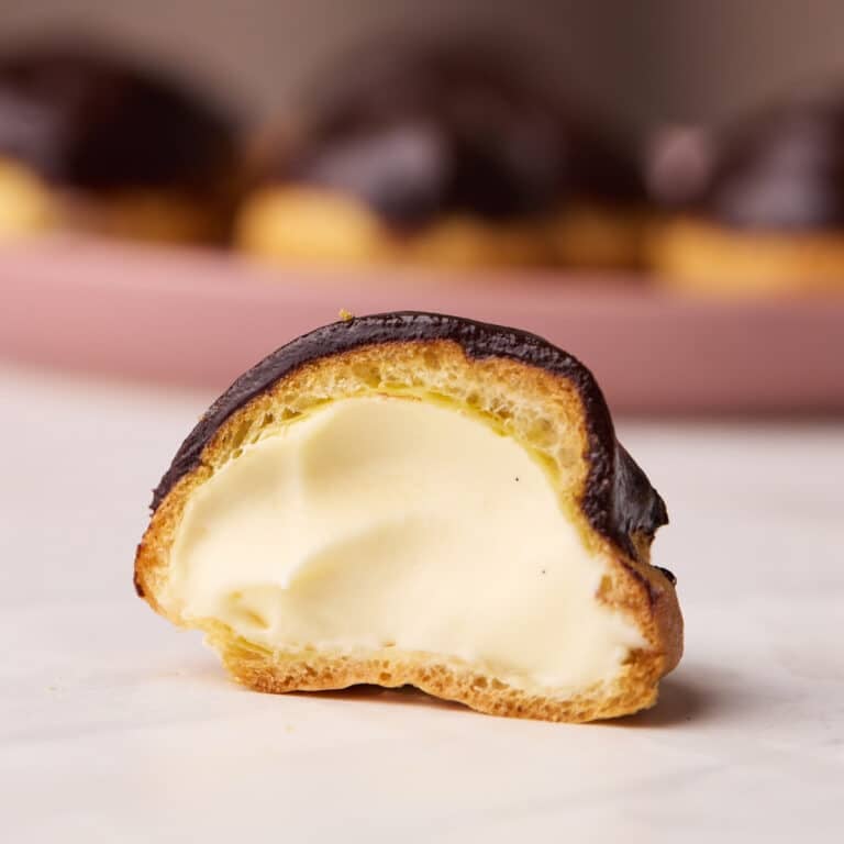 One profiterole cut in half to reveal the creamy pastry cream filling, a plate of more profiteroles are in the background on a pink plate.