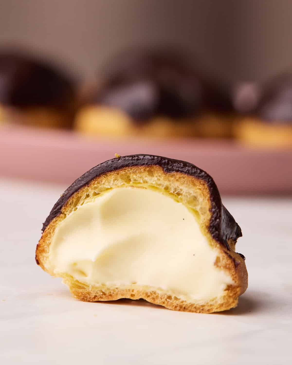 One profiterole cut in half to reveal the creamy pastry cream filling, a plate of more profiteroles are in the background on a pink plate.