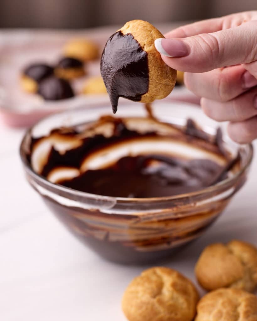 One profiterole being dipped into a bowl of warm chocolate glaze, the excess glaze is dripping off of the choux bun. 