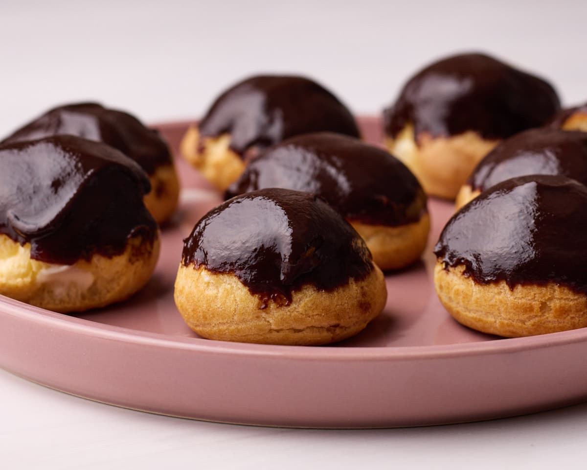 Homemade profiteroles with a shiny chocolate glaze, sitting on a pink plate.