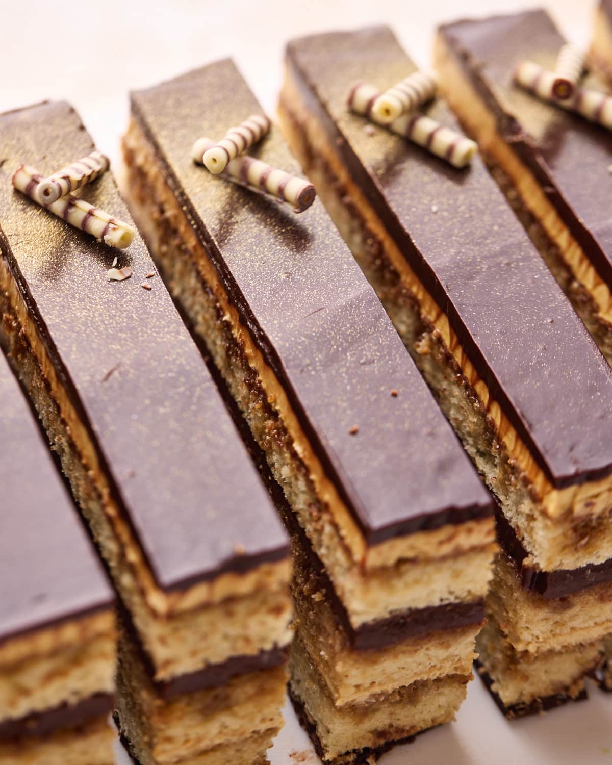 A row of opera cake slices with shiny chocolate glaze and edible glitter on top. 