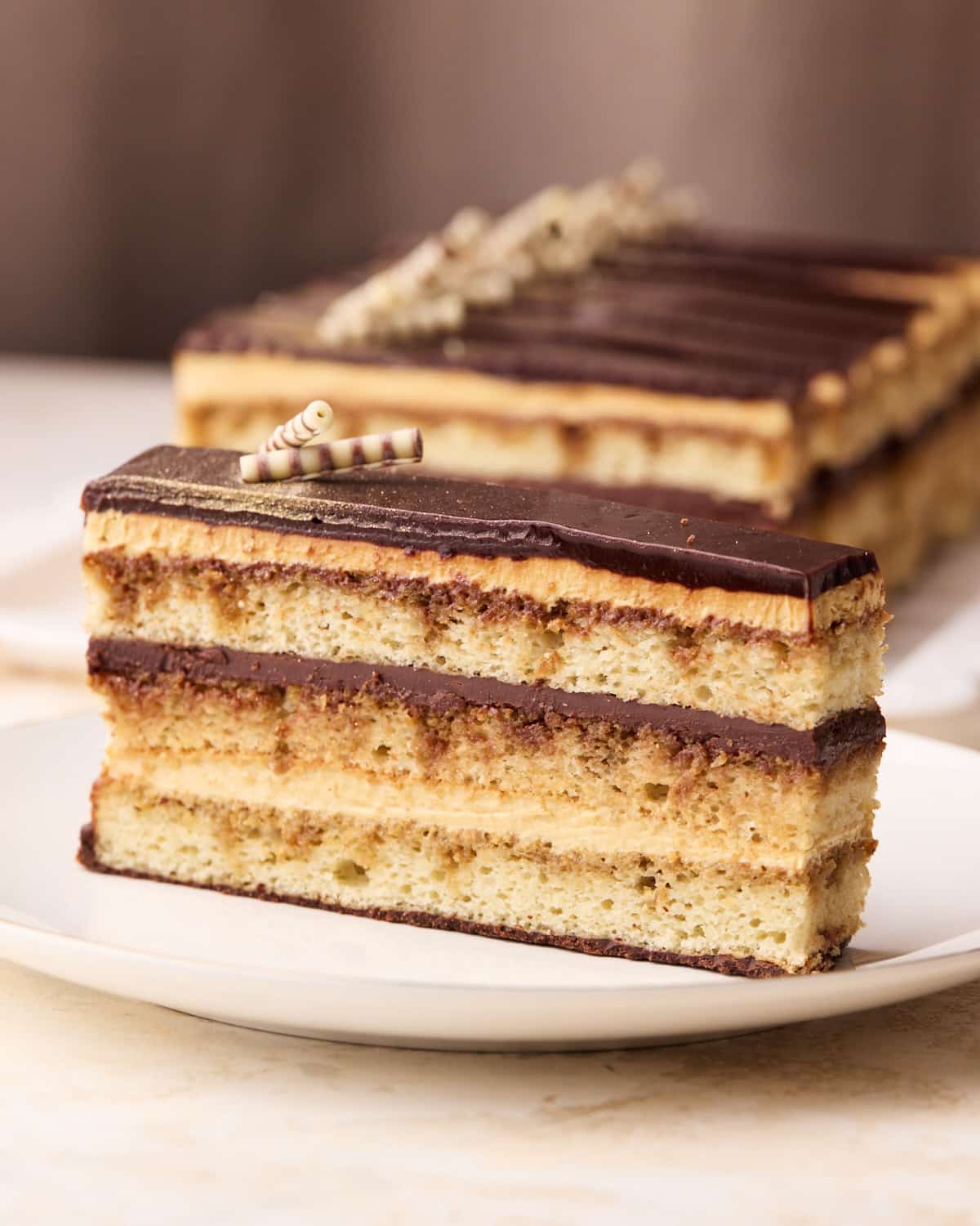 Slice of opera cake on a plate, showing all the beautiful layers, more slices are blurred out in the background. 