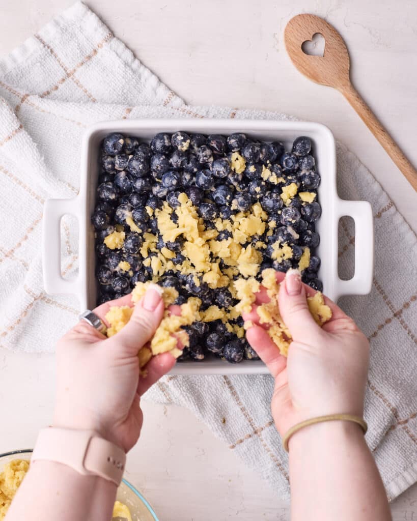 Sprinkling crumble topping over the top of fresh blueberries to make blueberry crumble.