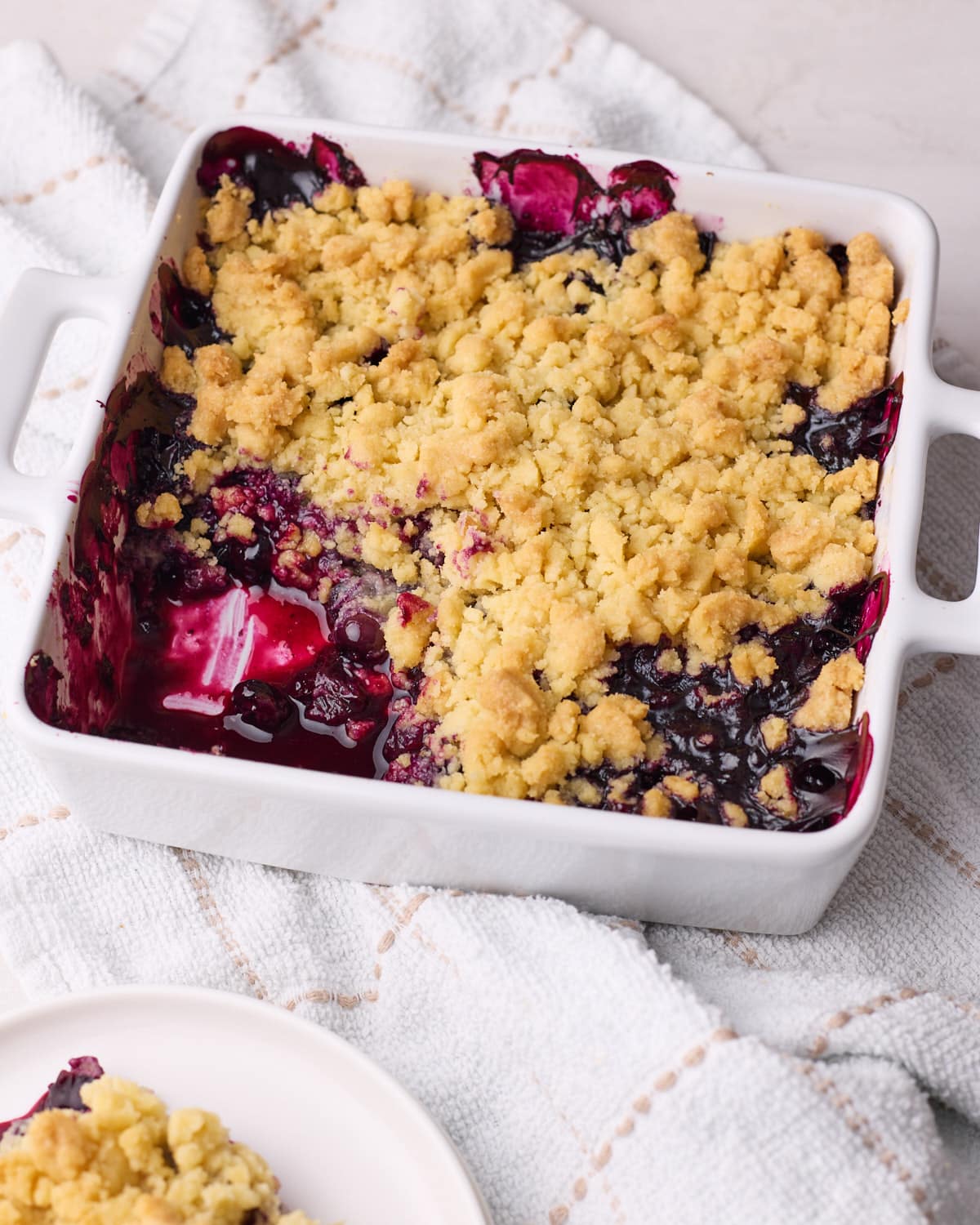 Homemade blueberry crumble in a baking dish with a portion scooped out of it to show the blueberry filling inside.