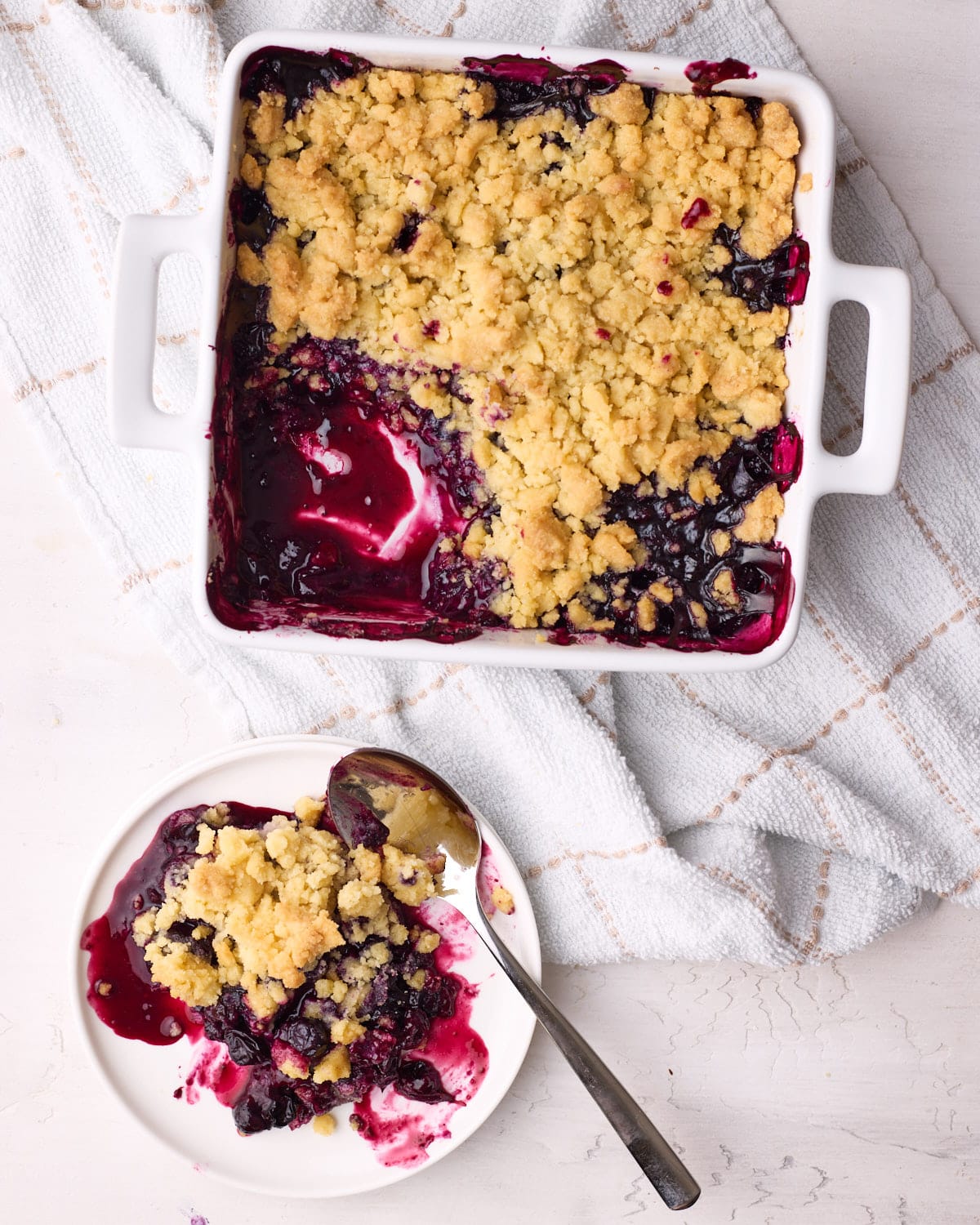 Blueberry crumble in a baking dish from above with a portion of it scooped out onto a small plate. 