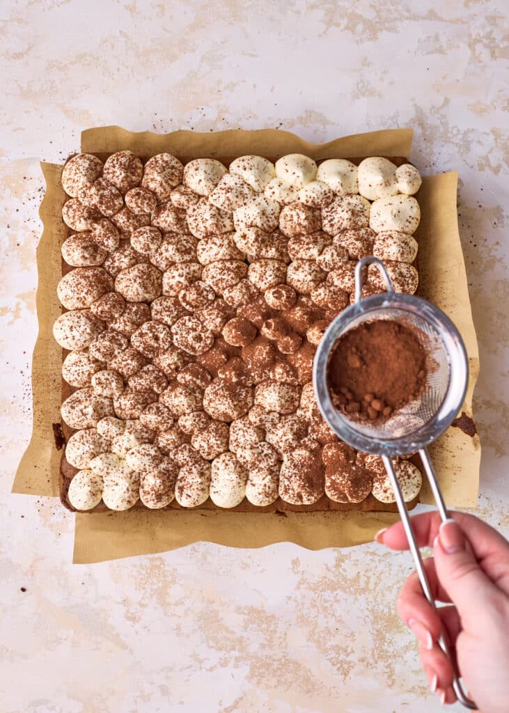 Dusting cocoa powder over the top of tiramisu brownies with a small metal sieve. 