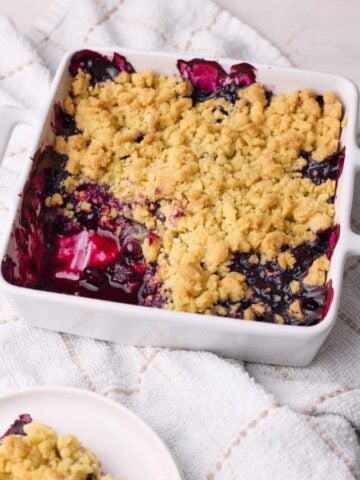 Homemade blueberry crumble in a baking dish with a portion scooped out of it to show the blueberry filling inside.