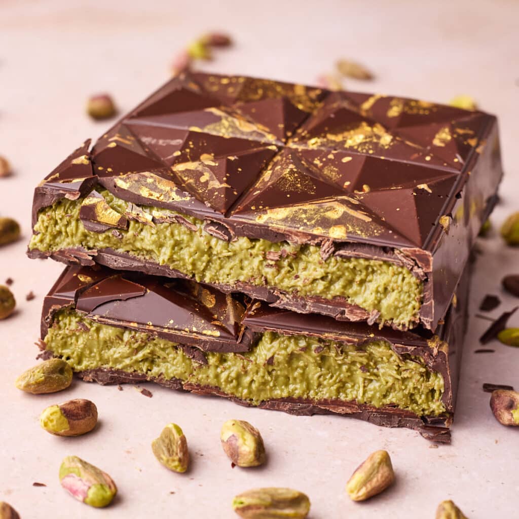Two Dubai Chocolate bars pictured with pistachio nuts around them - both bars cut in half to show the crunchy creamy pistachio filling.