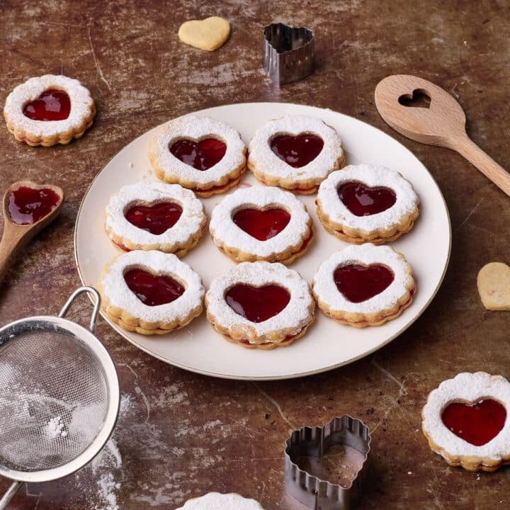Homemade linzer cookies on a plate, with a heart-shaped cut out window, surrounded by baking accessories.