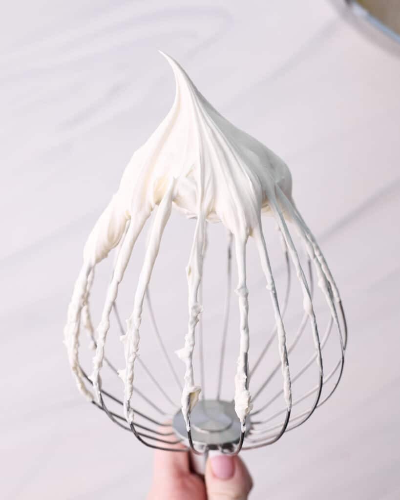 Whipped cream on a large whisk being held by a hand. 