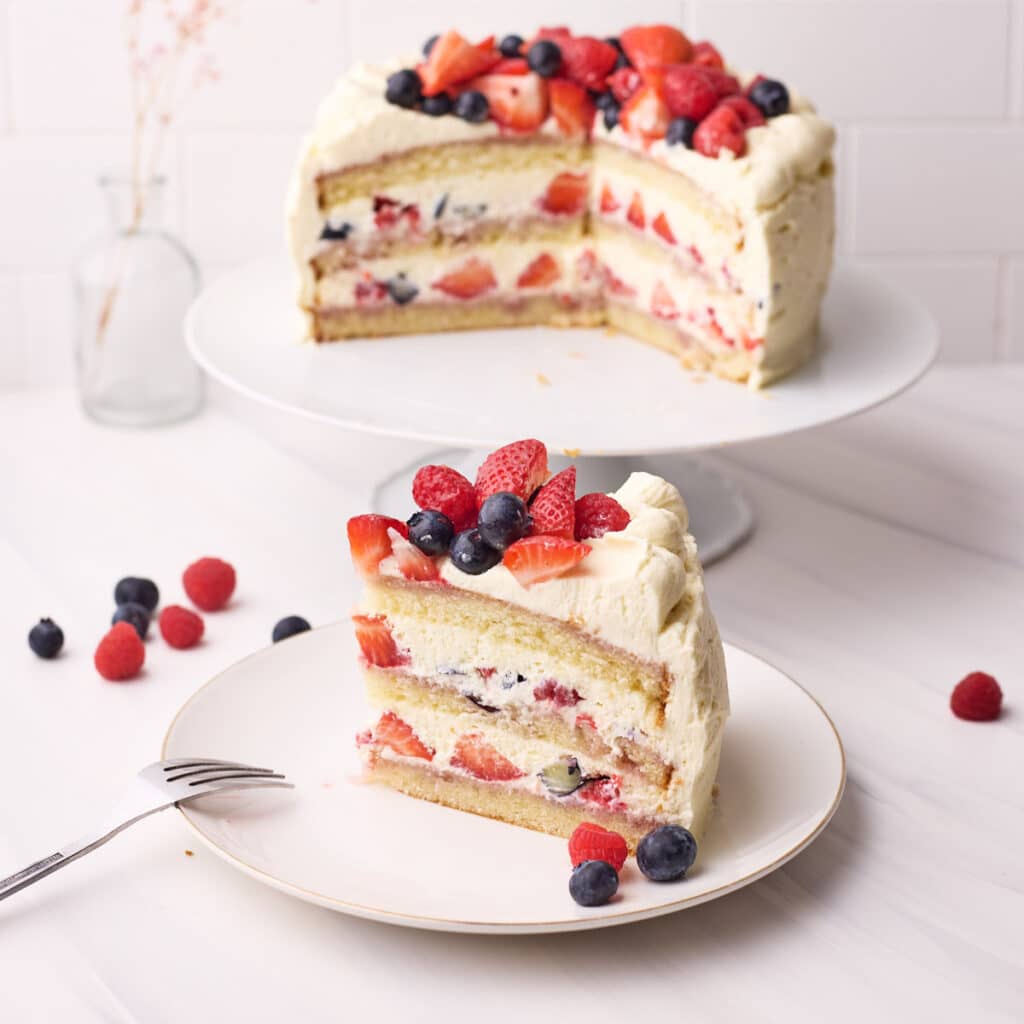 Slice of berry chantilly cake on a plate with the full cake in the background, fresh berries all around.