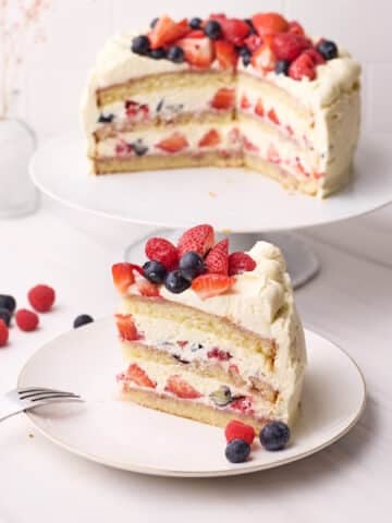 Slice of berry chantilly cake on a plate with the full cake in the background, fresh berries all around.