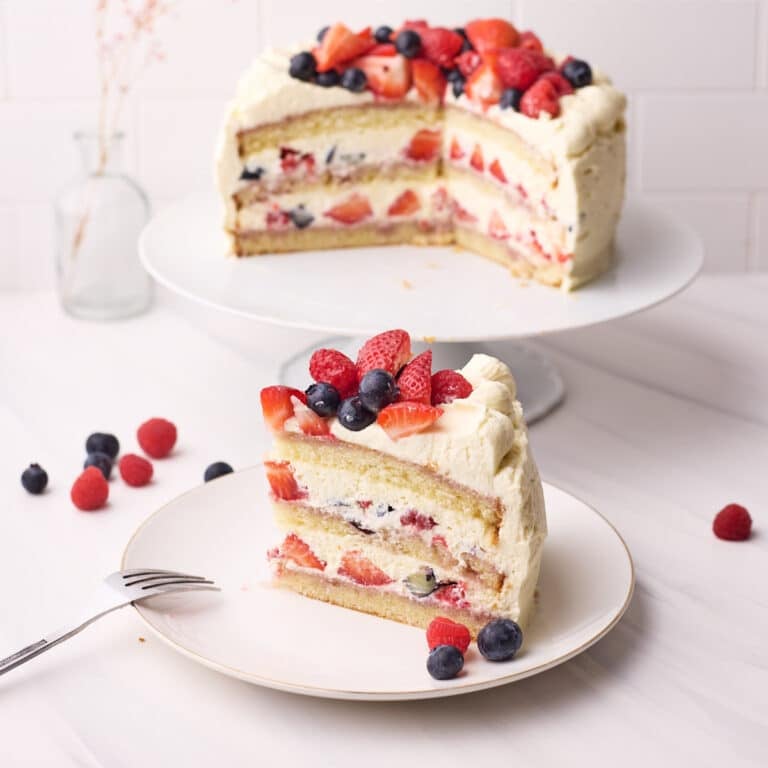 Slice of berry chantilly cake on a plate with the full cake in the background, fresh berries all around.