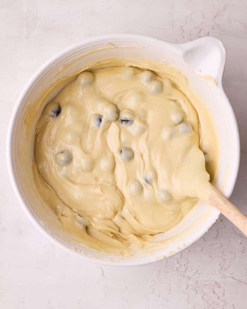 Blueberries being folded into the muffin batter in a large bowl with a wooden spoon.