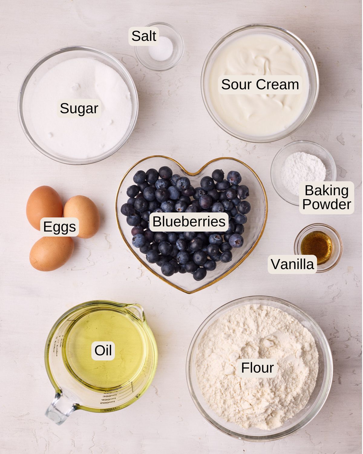 Labelled ingredients for blueberry muffins, including salt, sugar, sour cream, eggs, blueberries, baking powder, vanilla, vegetable oil, and flour.