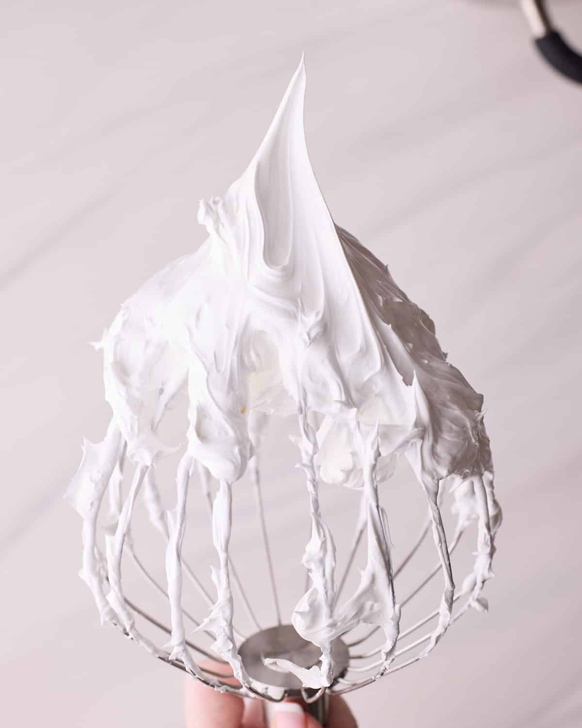 Meringue mixture being displayed on a whisk, showing the stiff peaks.