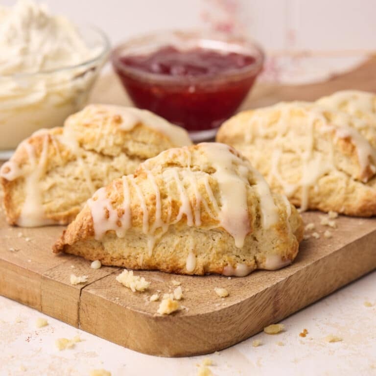 Scones on a wooden board drizzled with glaze, with jam and cream in bowls in the background.