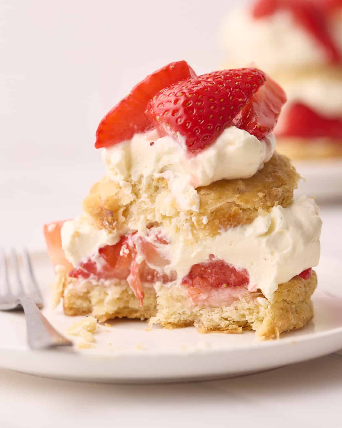 Close up of a half-eaten strawberry shortcake on a plate with a fork beside it.