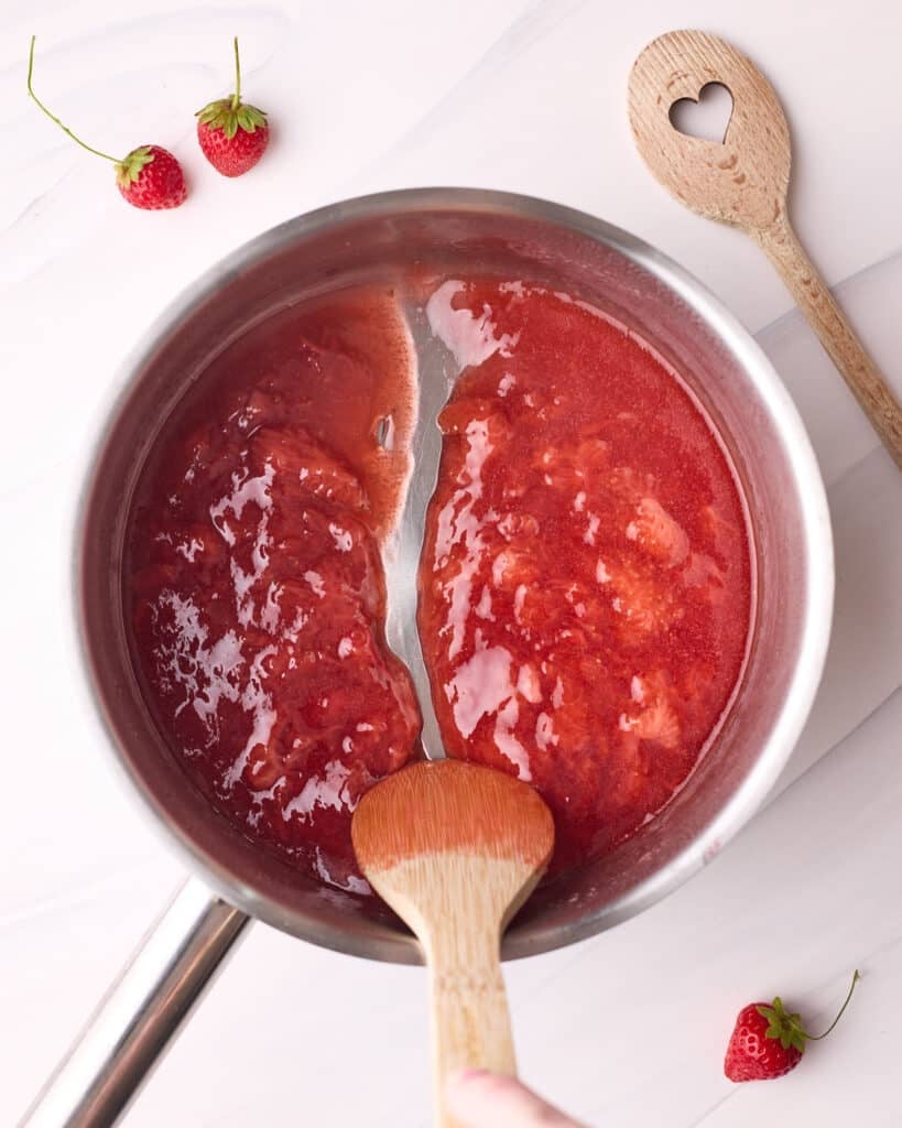 Strawberry glaze mixture in a saucepan after reaching the perfect consistency, which is being shown by a wooden spoon leaving a clear trail when stirring.