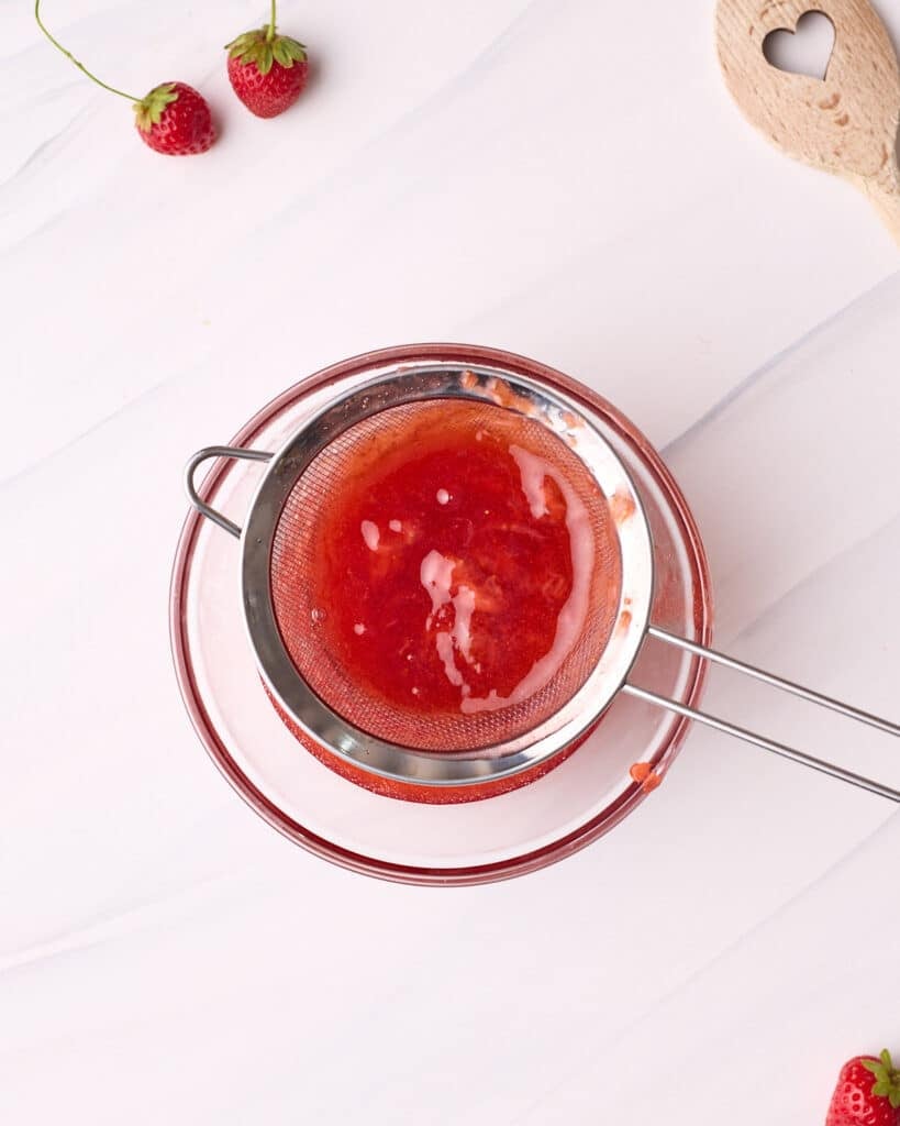 Strawberry glaze being strained through a fine mesh sieve into a small bowl.