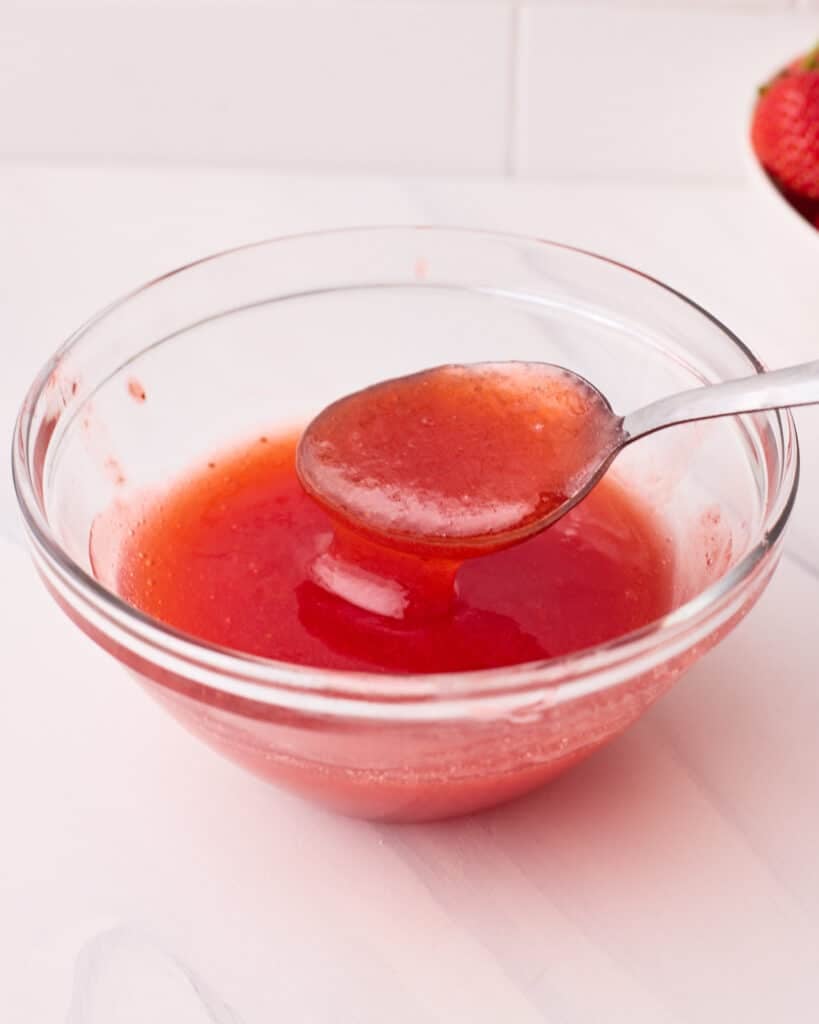 Strawberry glaze in small glass bowl being spooned out with a teaspoon.
