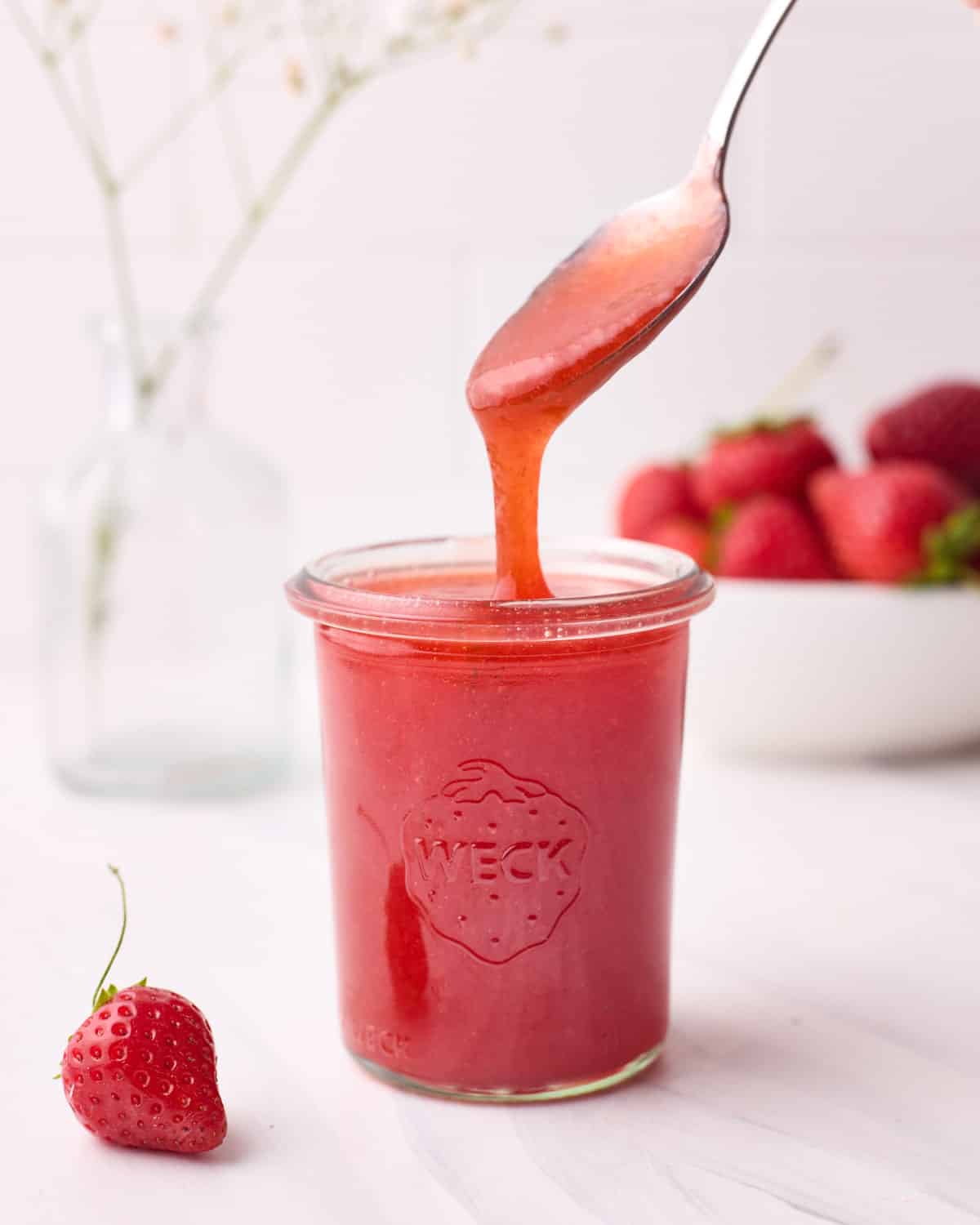 Strawberry glaze in a mason jar, with a spoonful of glaze being dripped into it.
