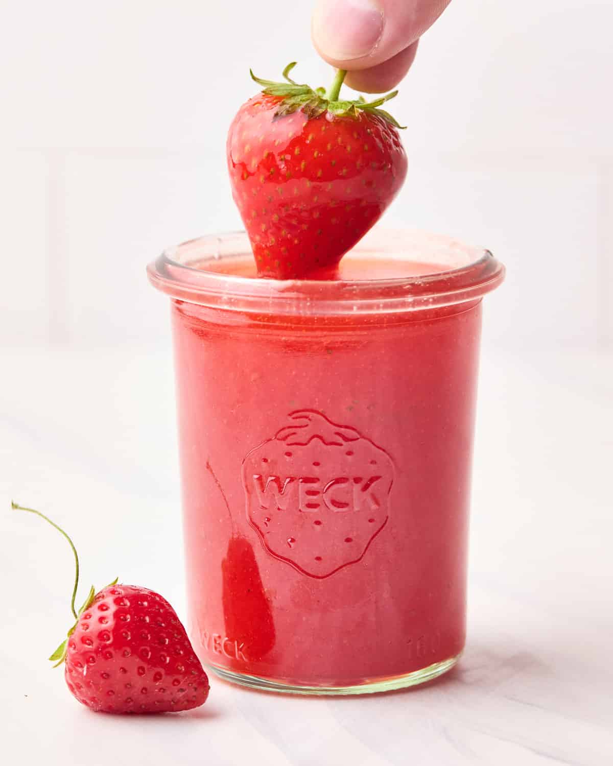 Strawberry glaze in a mason jar with a strawberry being dipped in it.