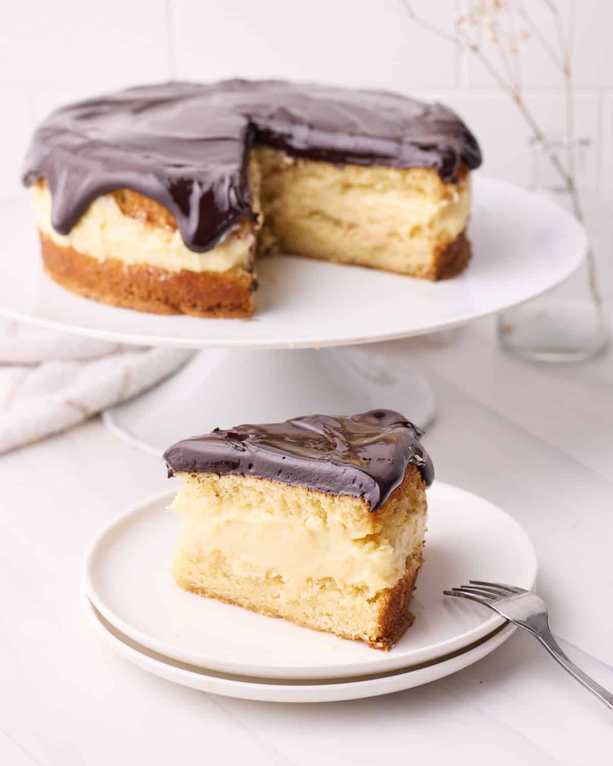 Slice of Boston cream pie on a plate with the full cake in the background - the distinct layers of sponge cake, pastry cream filling and chocolate glaze are visible.