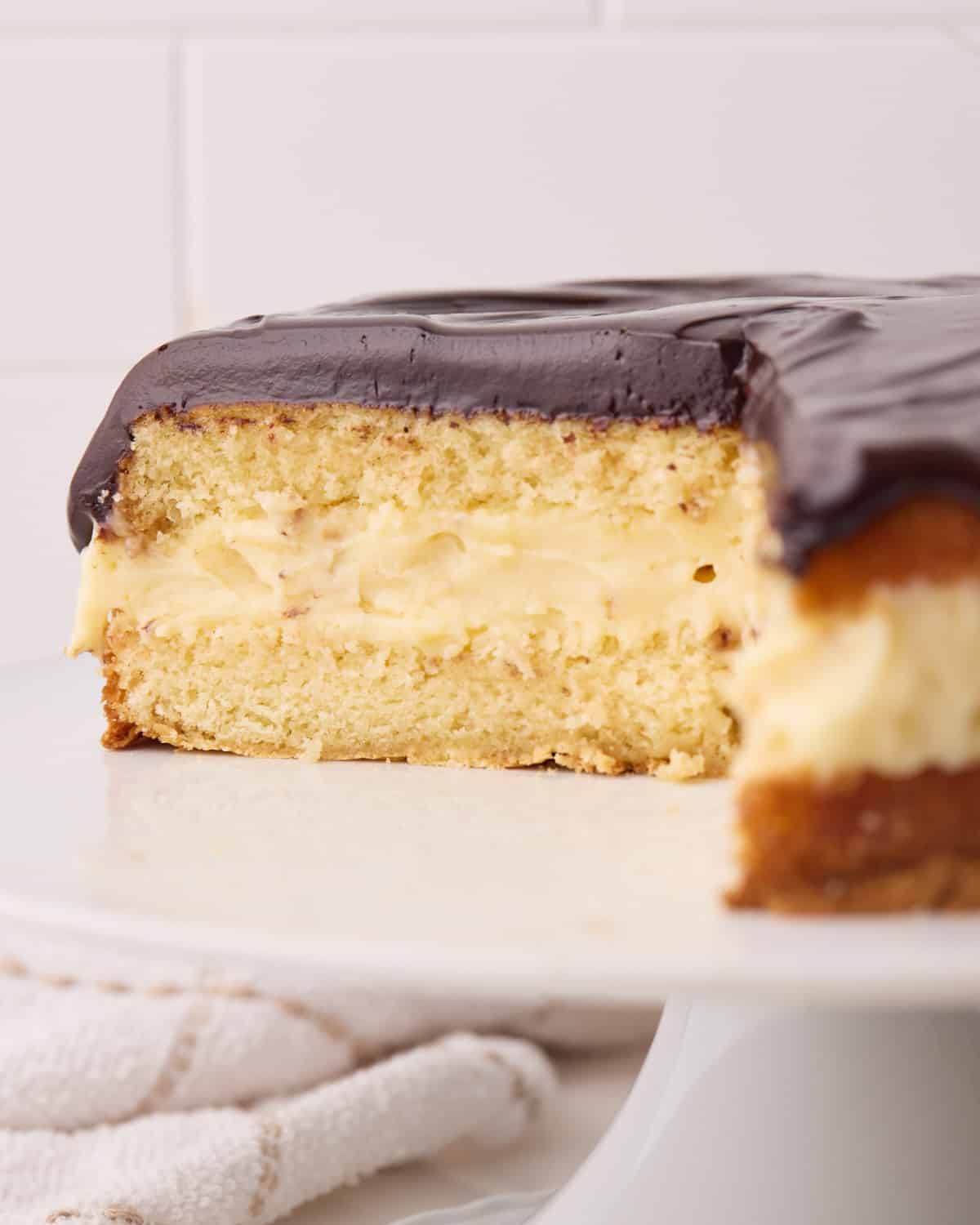 Side shot of a Boston cream pie with a slice taken out of it to reveal the fluffy vanilla cake, custard filling and shiny chocolate glaze.